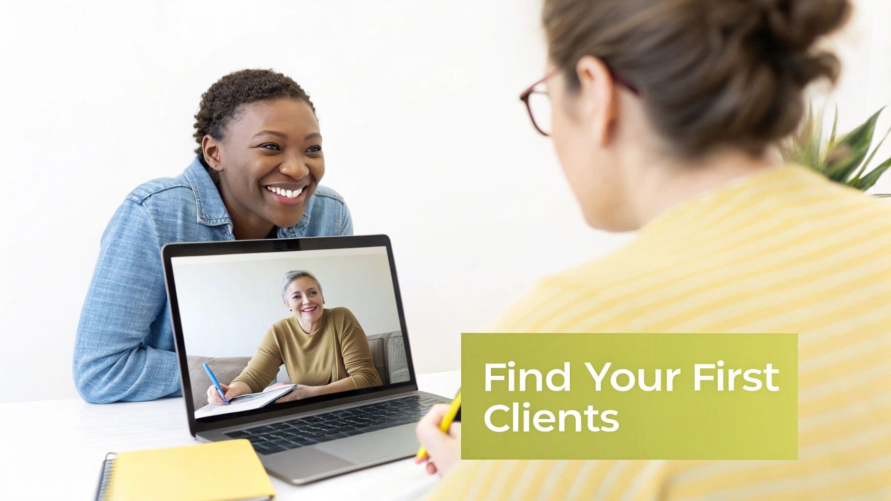 Two smiling women in a virtual coaching session, one looking at a laptop, the other taking notes.